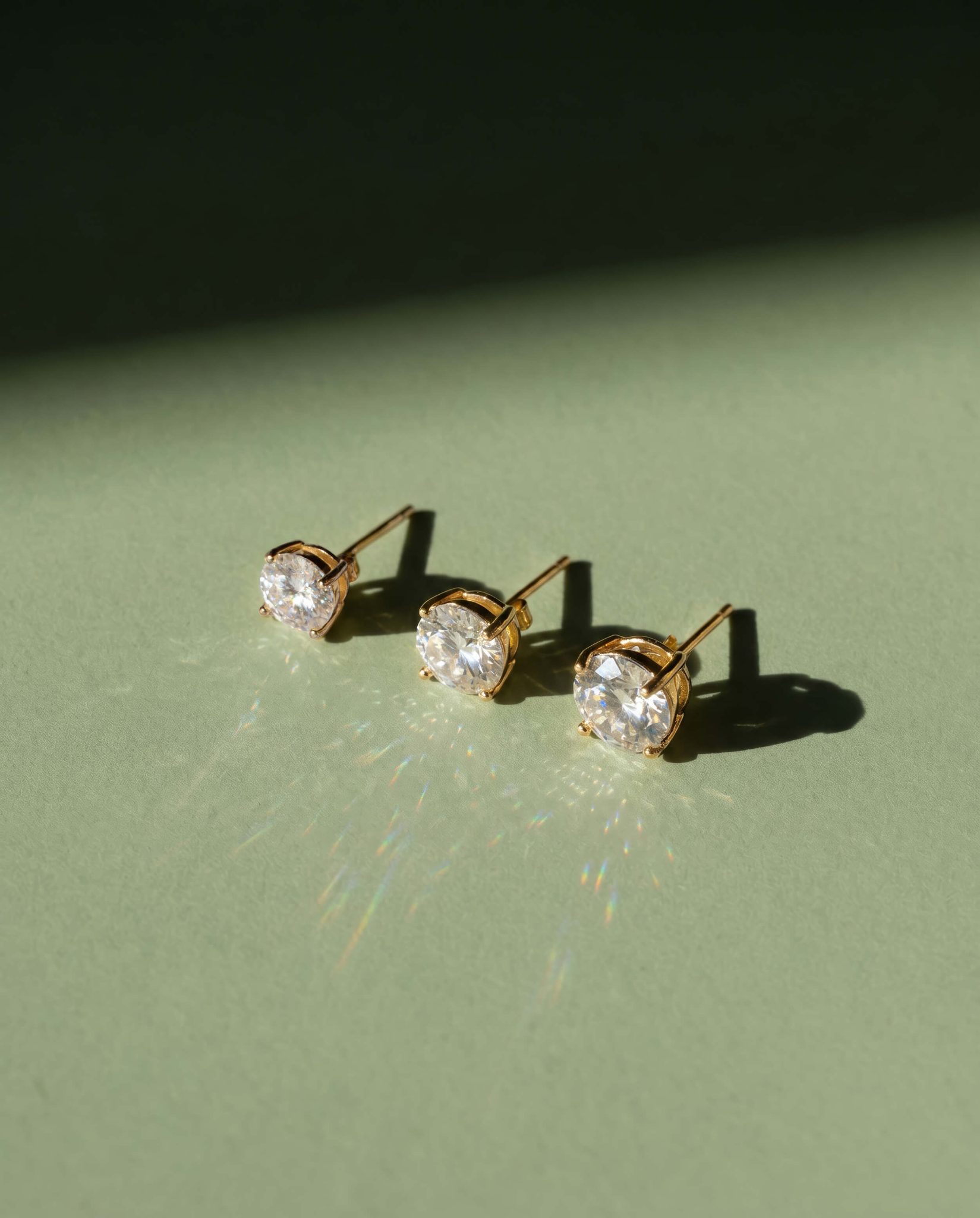 Three crystal stud earrings in different sizes photographed on sage green background with directional light showing size through shadow and light refraction — commercial jewelry photography