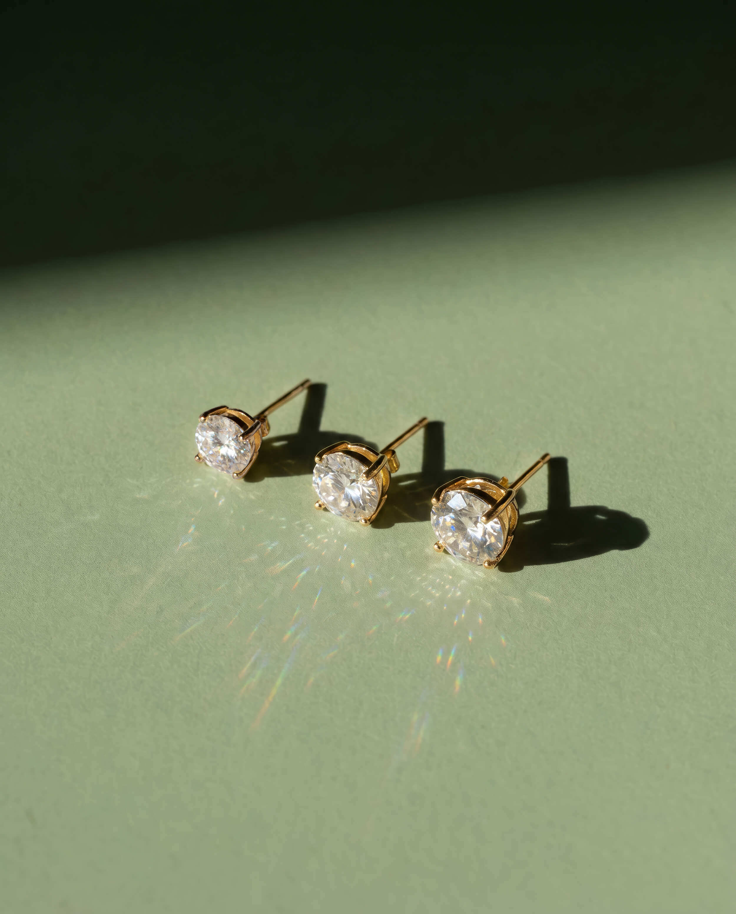 Three crystal stud earrings in different sizes photographed on sage green background with directional light showing size through shadow and light refraction — commercial jewelry photography